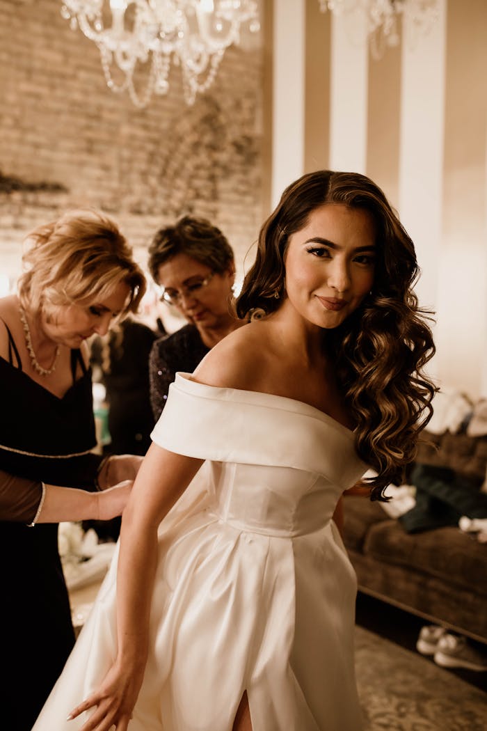 Home A bride with long wavy hair in a stunning white dress, preparing for her wedding with assistants indoors.