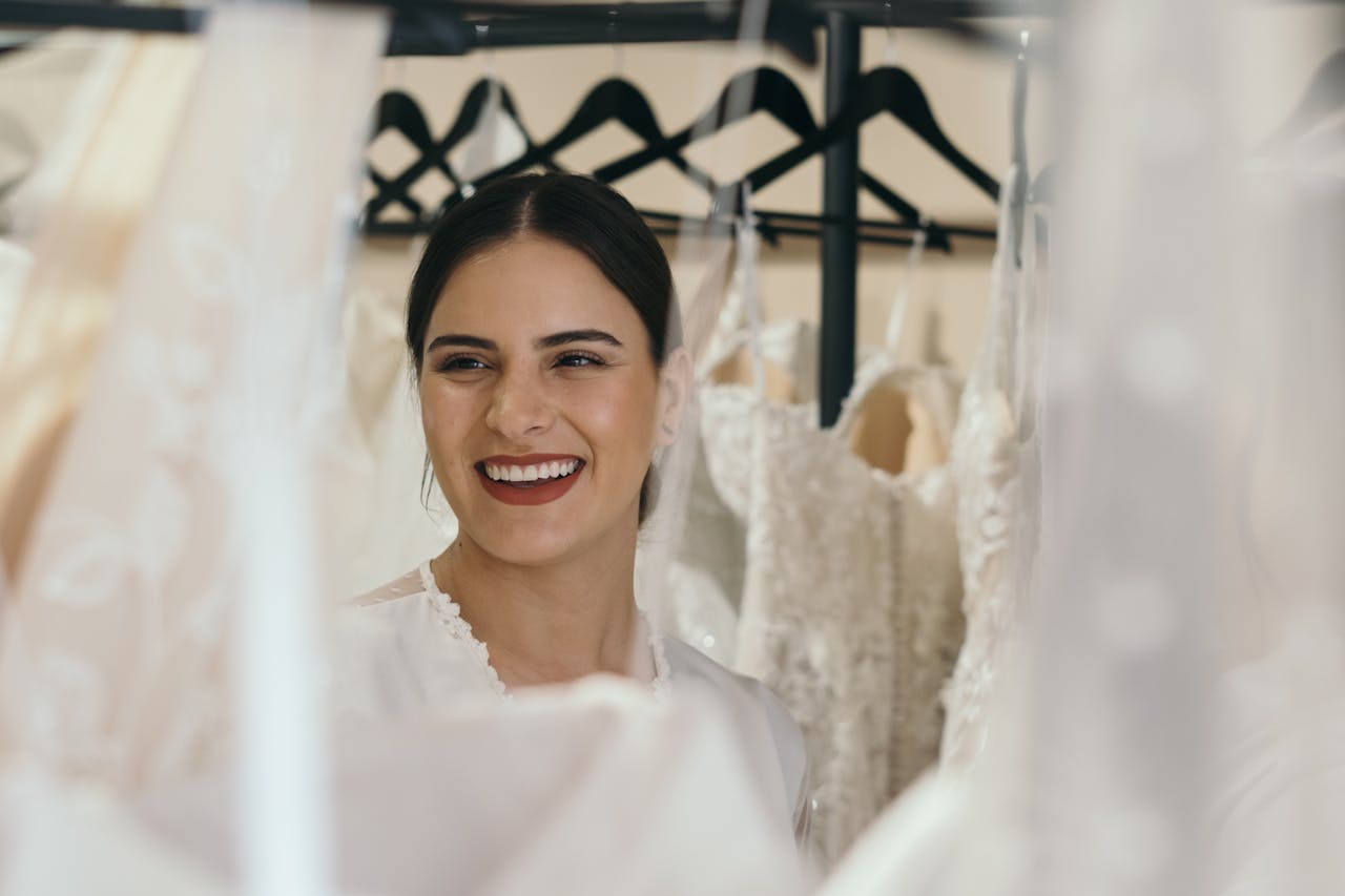 Home A joyful bride smiles while surrounded by beautiful wedding dresses on hangers.